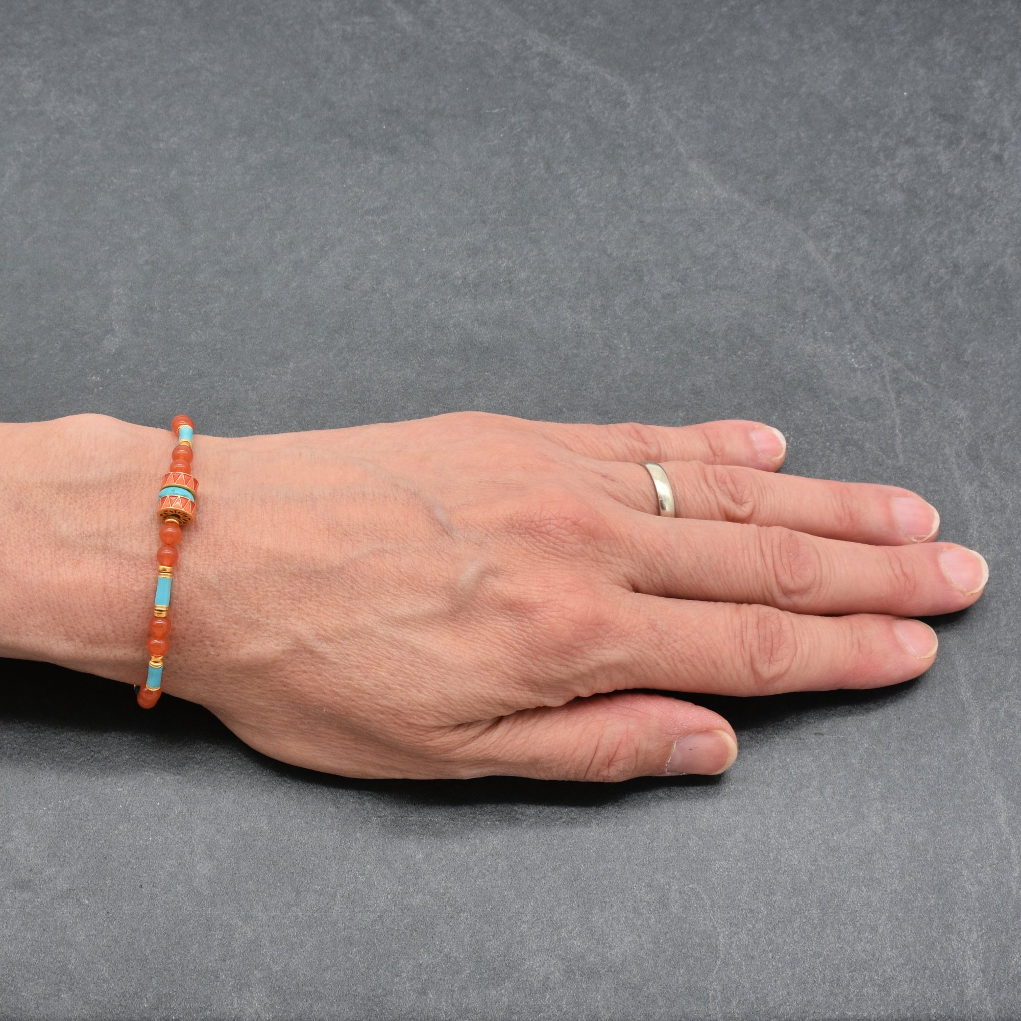 A beaded bracelet with orange and turquoise beads worn on a person's wrist, shown against a dark gray slate background. The person's hand is resting flat on the surface, and they are wearing a simple gold band on their ring finger - handmade with love in Colorado by Ruby Acorn.