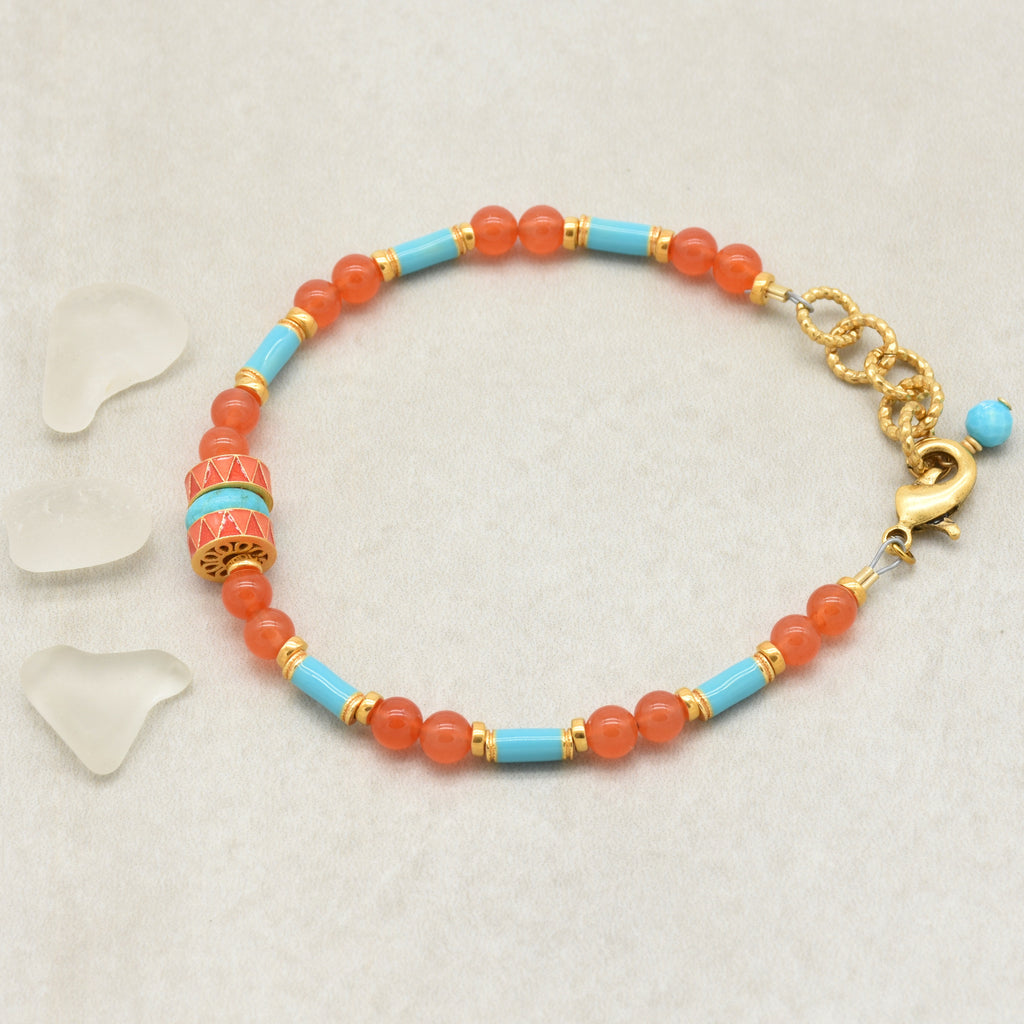 A beaded bracelet with round coral-orange beads, turquoise-blue cylindrical beads, and a decorative coral-and-gold spacer bead, displayed on a light background with small clear stone chips - handmade with love in Colorado by Ruby Acorn.