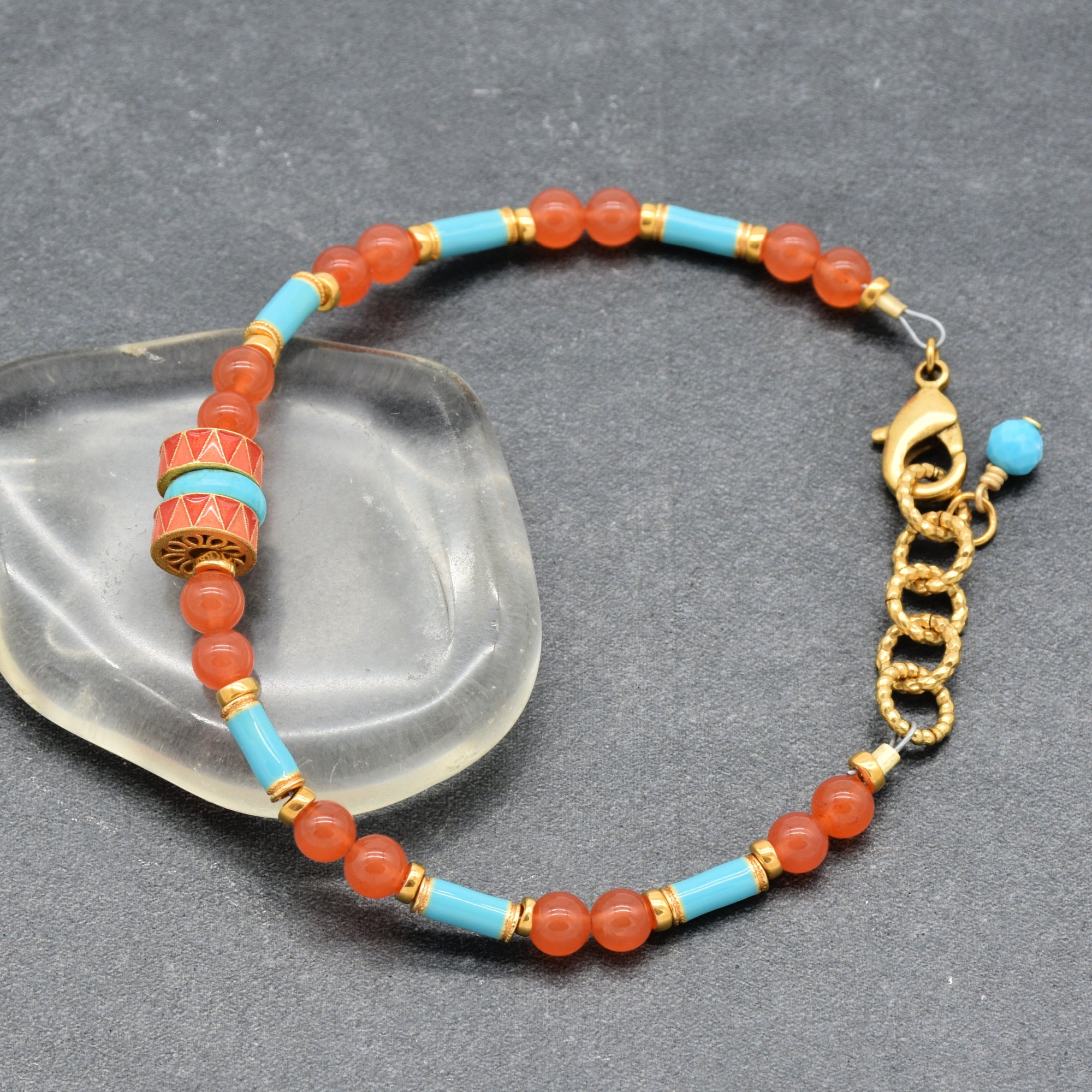 A beaded bracelet with coral pink beads, light blue turquoise beads, and gold-toned spacer rings, featuring a decorative cylindrical accent bead and a gold-toned lobster clasp with a small blue bead charm, displayed on a dark gray textured surface with a clear glass prop - handmade with love in Colorado by Ruby Acorn.