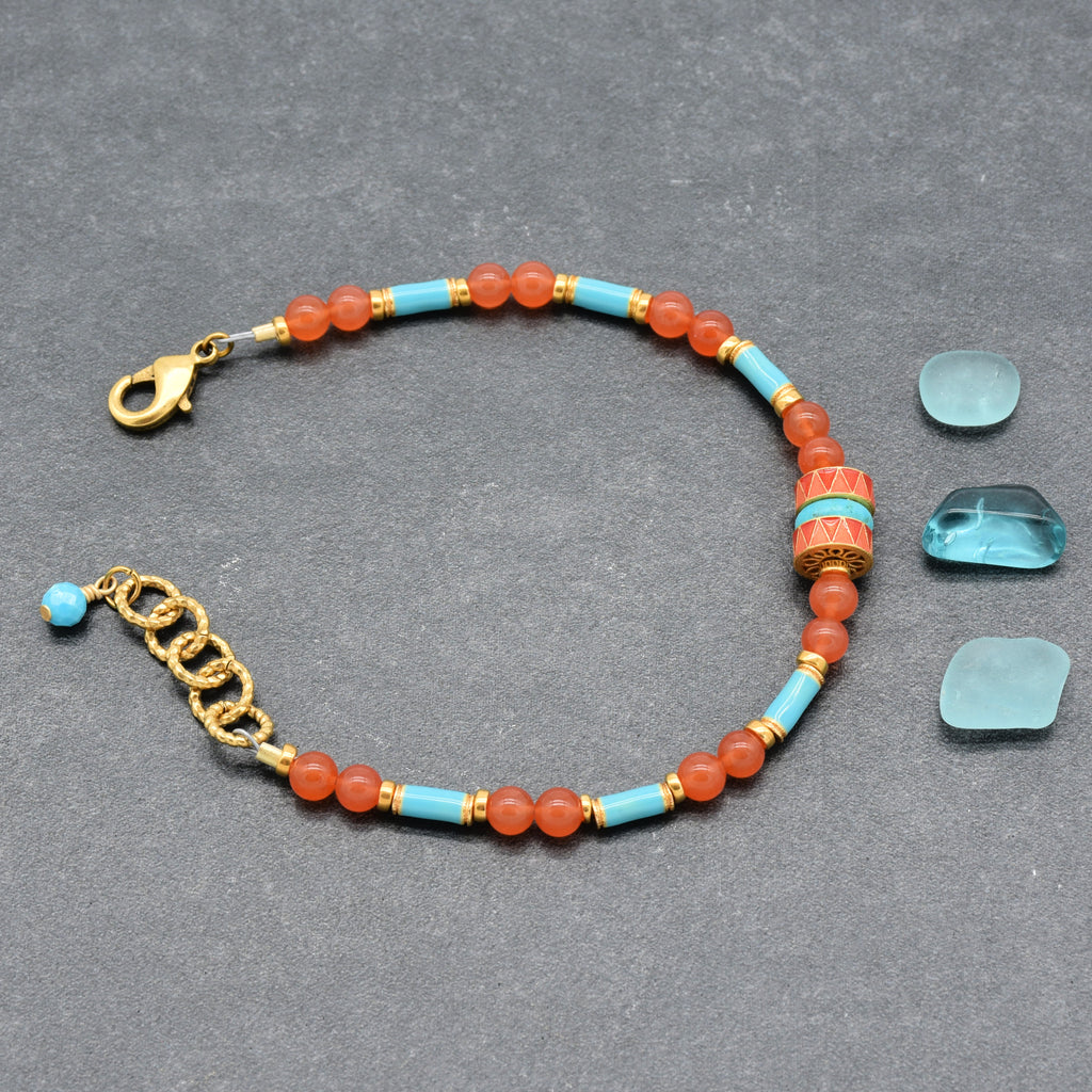 A beaded bracelet featuring round coral-orange beads, light blue turquoise beads, and gold spacer rings, with a central decorative cylindrical bead and a gold clasp, shown on a gray background - made with love in Colorado by Ruby Acorn.