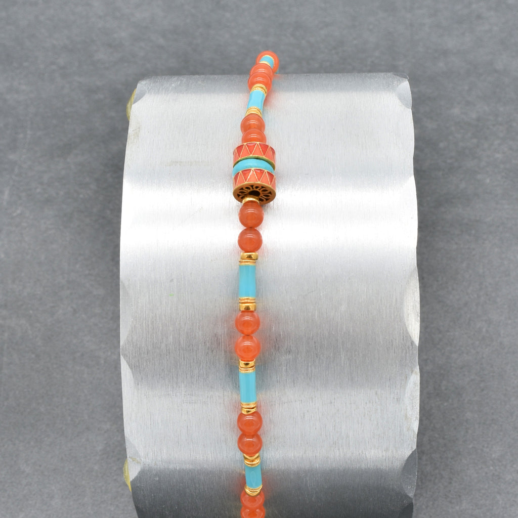 A beaded bracelet featuring round coral-orange beads, light blue turquoise beads, and gold spacer rings, with a central decorative cylindrical bead and a gold clasp, shown on a silver display on a gray background - made with love in Colorado by Ruby Acorn.