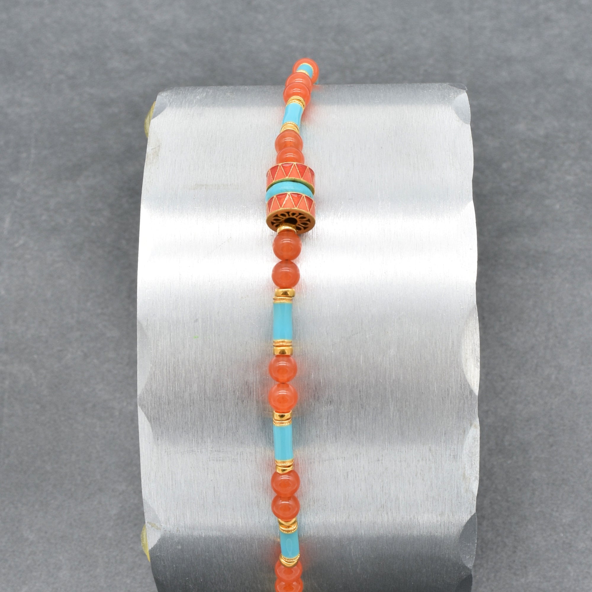 A beaded bracelet featuring round coral-orange beads, light blue turquoise beads, and gold spacer rings, with a central decorative cylindrical bead and a gold clasp, shown on a silver display on a gray background - made with love in Colorado by Ruby Acorn.