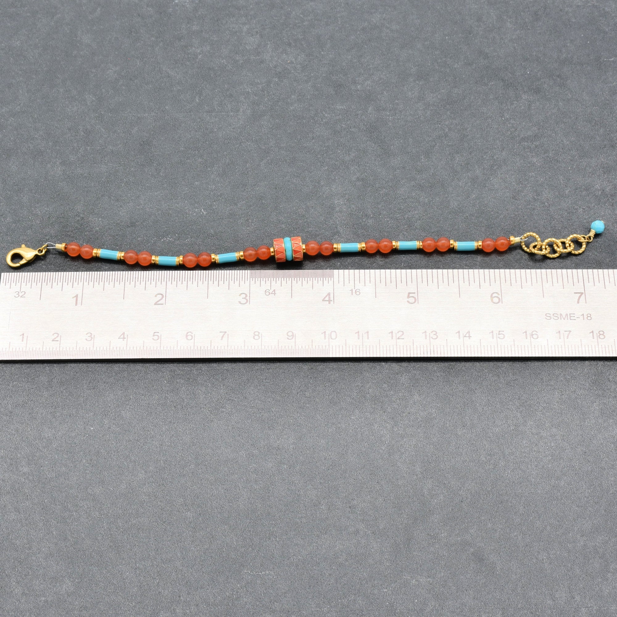 A beaded bracelet with orange-red and turquoise blue beads, gold-tone clasp, and extender chain, shown on a gray background with a ruler for scale - handmade with love in Colorado by Ruby Acorn.