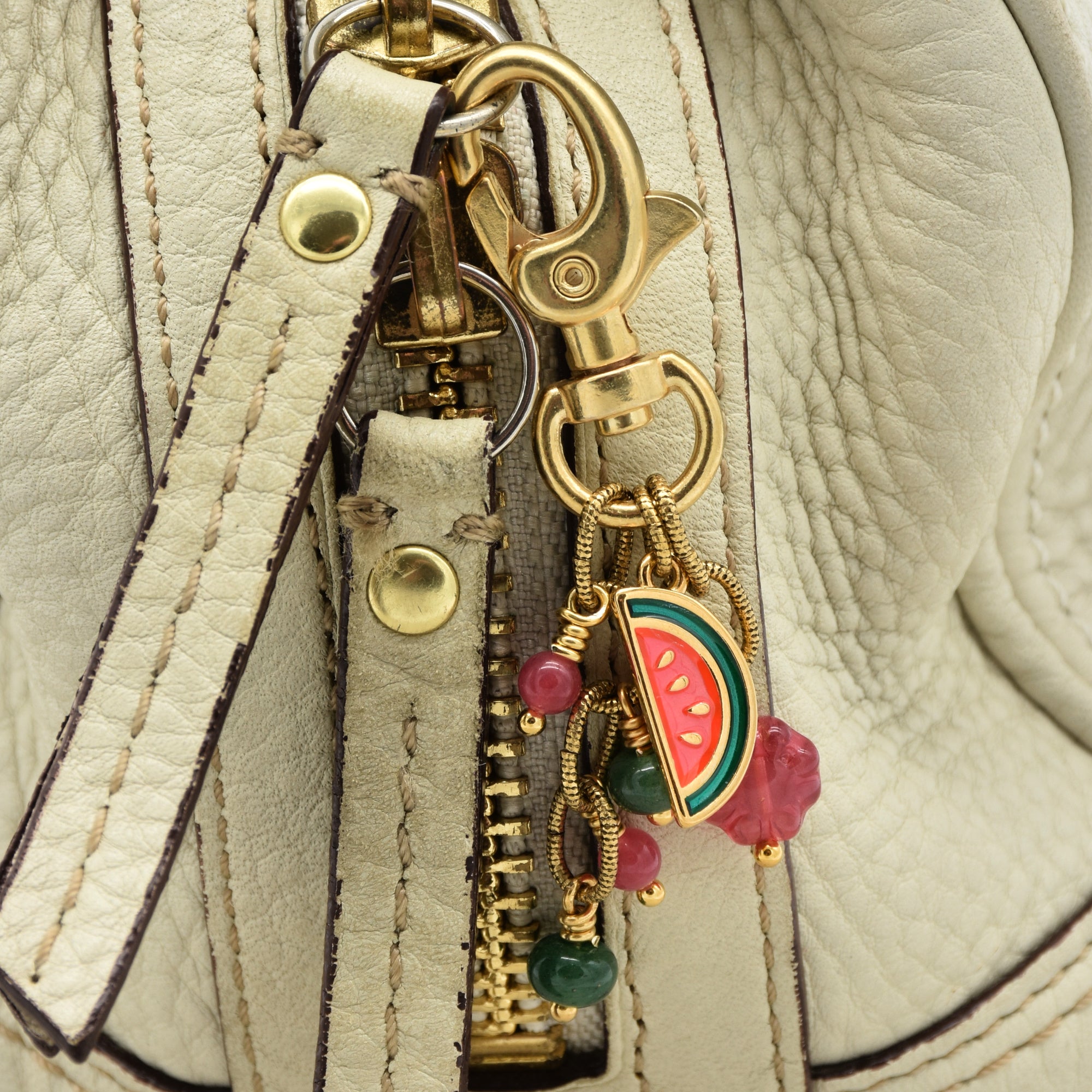 Close-up of a cream-colored textured leather bag featuring a gold-tone zipper pull with a gold swivel clasp and colorful charms, including a watermelon slice and pink beads - made with love by Ruby Acorn.