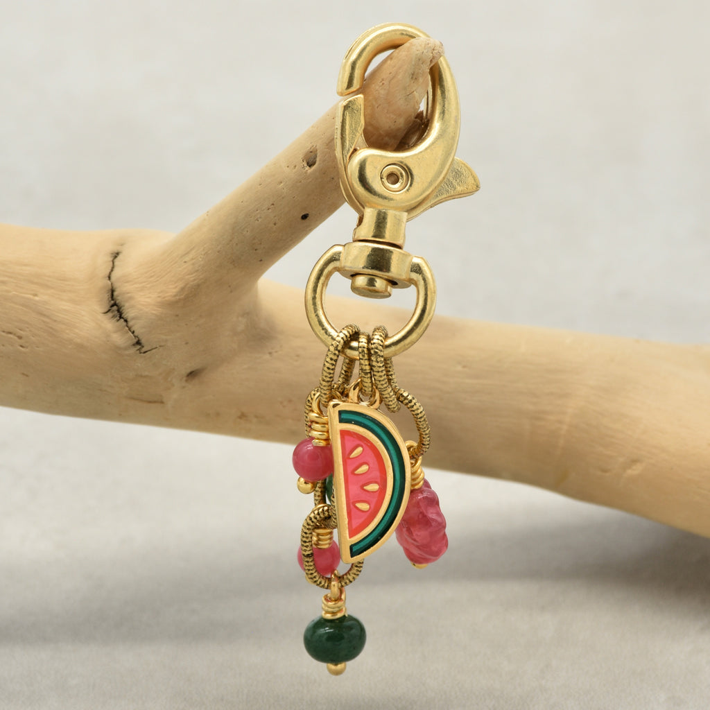 A gold-toned lobster clasp keychain charm featuring a watermelon slice charm, pink beads, and a green bead, displayed on a wooden branch against a light gray background - made with love by Ruby Acorn.