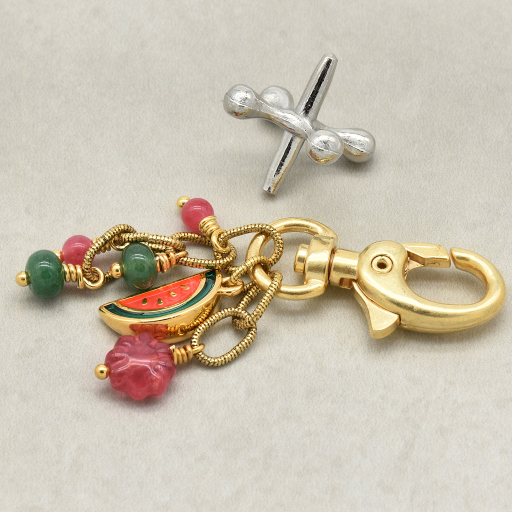 A gold-toned keychain or bag charm with a lobster clasp, twisted chain links, pink and green beads, a watermelon slice charm, and a pink flower charm, shown on a light neutral background - made with love by Ruby Acorn.