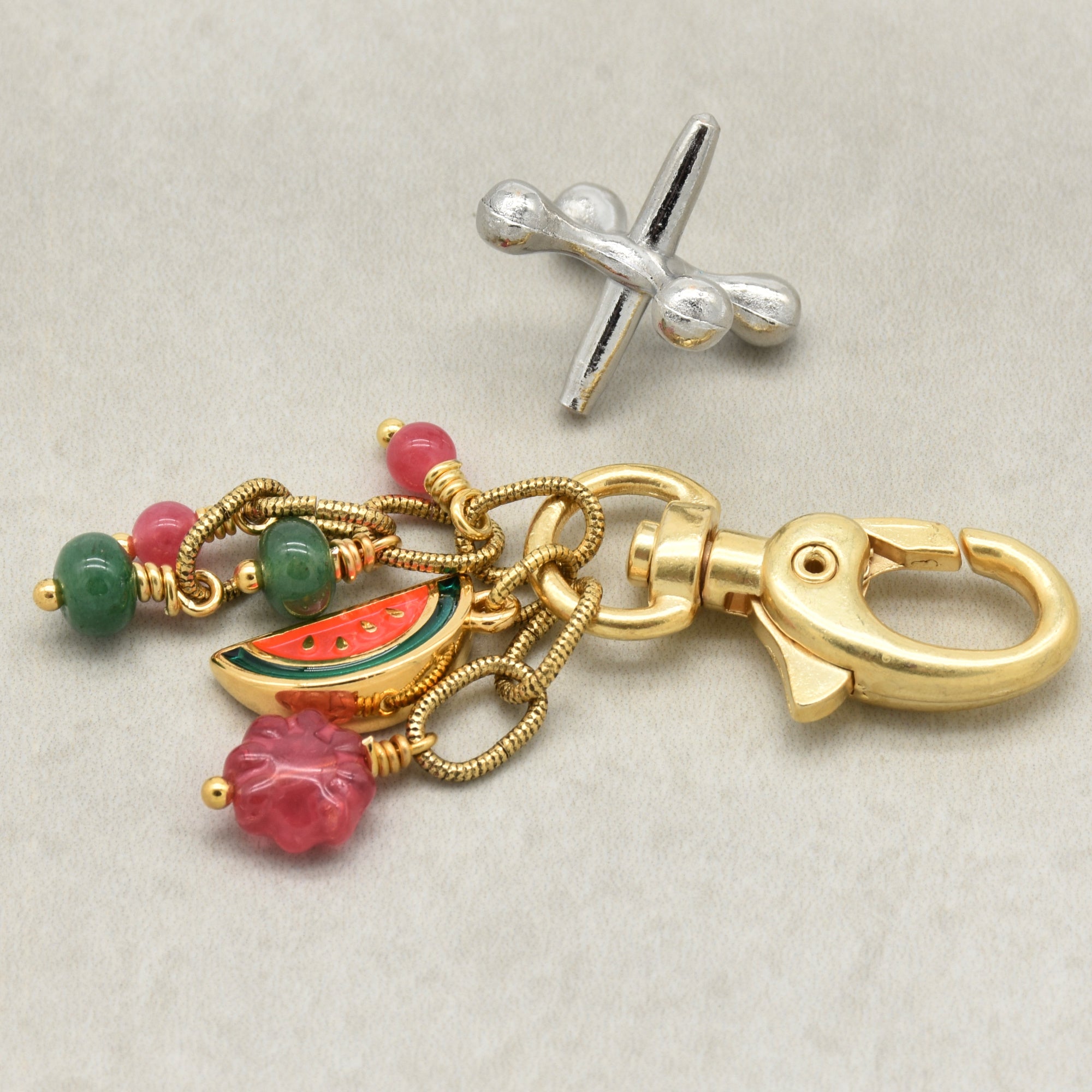 A gold-toned keychain or bag charm with a lobster clasp, twisted chain links, pink and green beads, a watermelon slice charm, and a pink flower charm, shown on a light neutral background - made with love by Ruby Acorn.