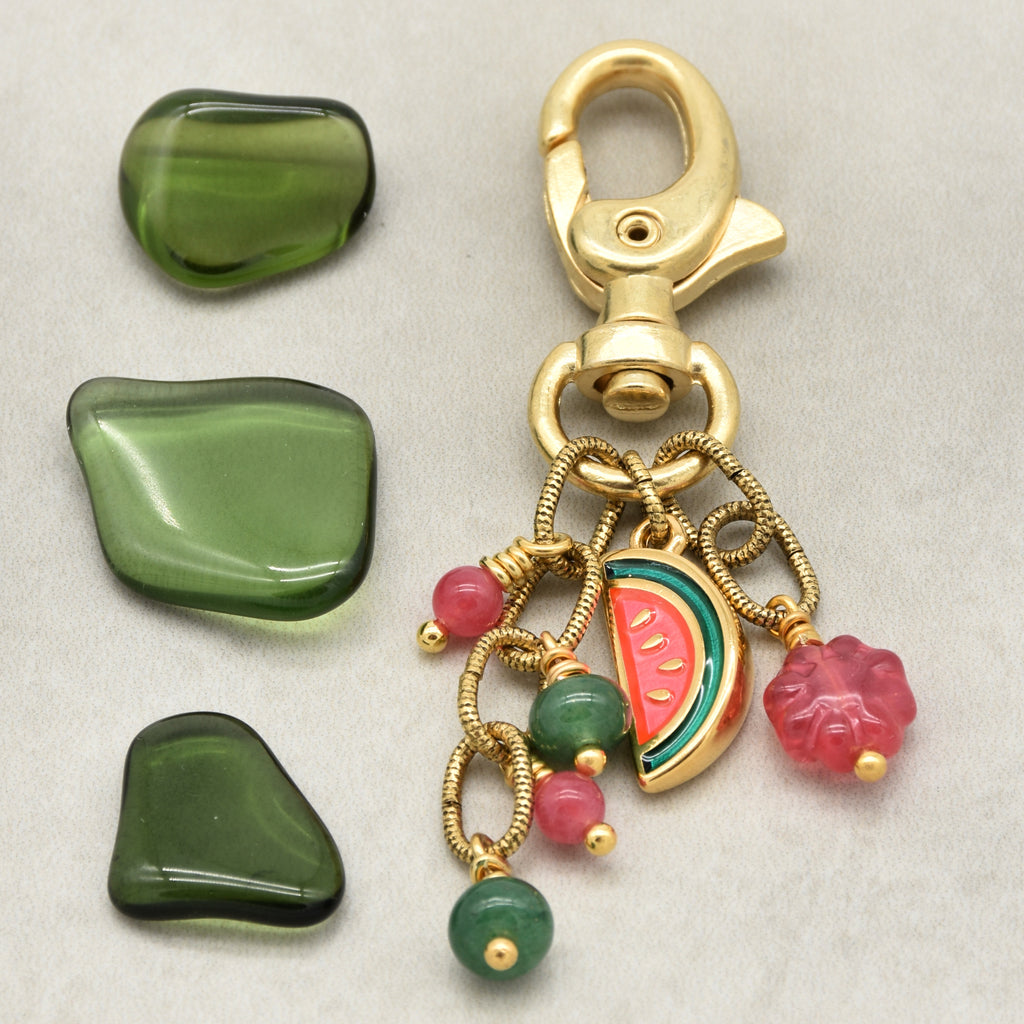 A gold-toned keychain or bag charm with a lobster clasp and dangling charms, shown alongside three smooth green stone pieces on a light gray background. The charm includes a watermelon slice charm, pink flower charms, green and pink beads, and a coiled wire detail - made with love by Ruby Acorn.