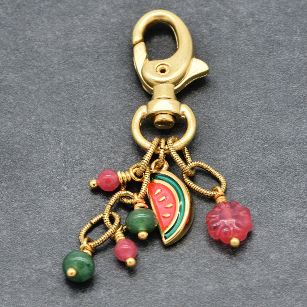 A gold-tone lobster clasp keychain charm featuring dangling beads and charms, including a watermelon slice and pink and green beads, set against a dark gray textured background - made with love by Ruby Acorn.