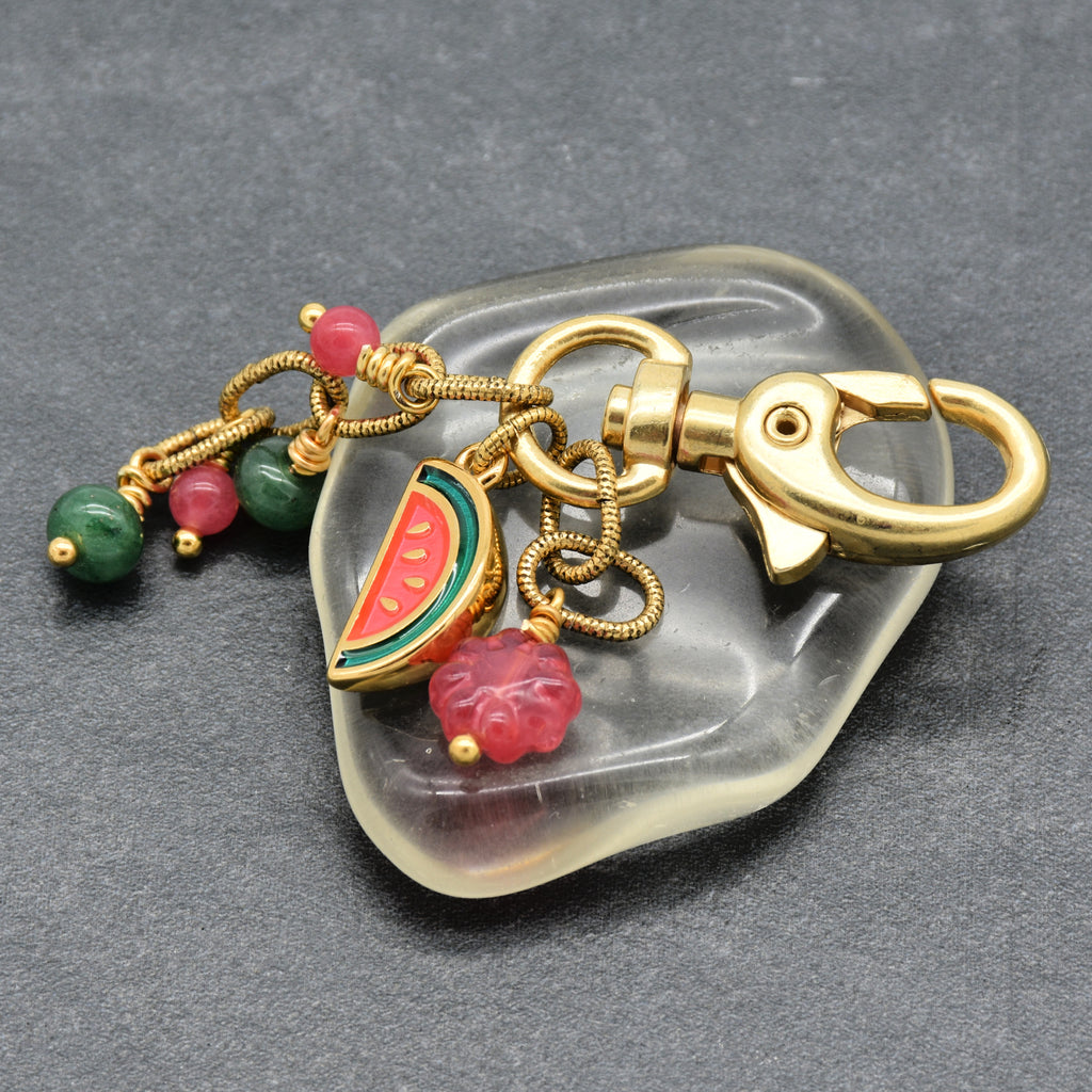 A gold-tone metal keychain with a lobster clasp, featuring dangling charms including a watermelon slice, a pink flower, green and pink beads, and a small pink charm, displayed on a clear acrylic stand over a dark gray textured background - made with love by Ruby Acorn.
