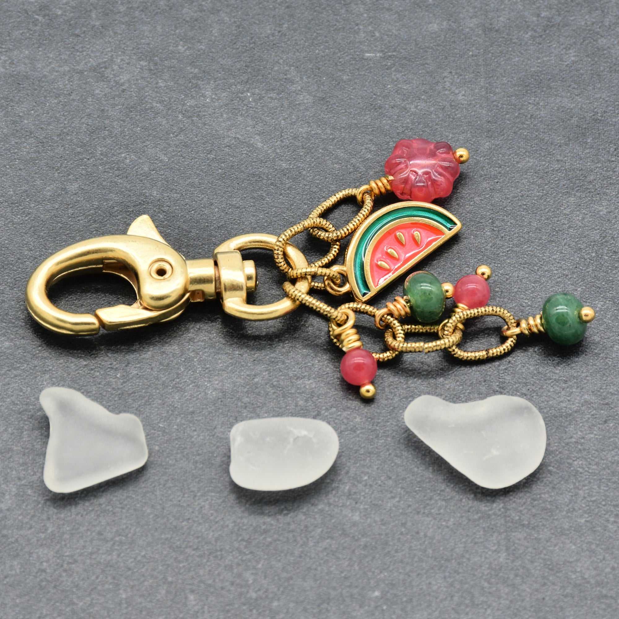 A gold-tone lobster clasp keychain or bag charm with a watermelon slice charm, pink and green beads displayed below on a dark gray textured background - made with love by Ruby Acorn.