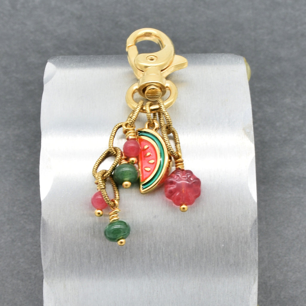A gold-toned lobster clasp keychain or bag charm with dangling charms, including a watermelon slice and pink flower beads, displayed on a white cylindrical stand against a gray background - made with love by Ruby Acorn.