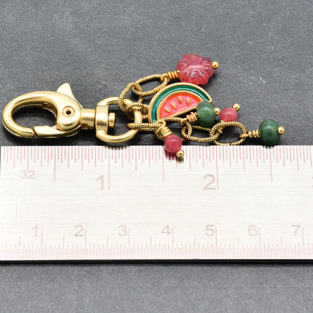 A gold-tone metal keychain or bag charm with a lobster clasp, featuring a watermelon slice charm, green and pink beads, and a small pink flower charm, shown above a ruler for scale on a dark gray background - made with love by Ruby Acorn.