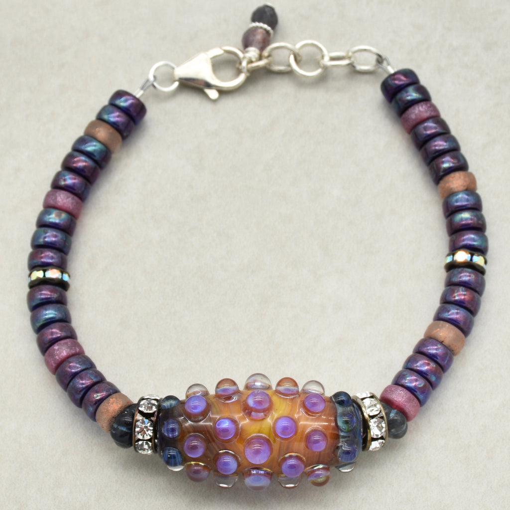 This image shows a bracelet. It features a central decorative bead with raised purple dots, surrounded by smaller beads in shades of purple, blue, and pink, and it has a clasp for fastening. The size and design are consistent with a bracelet.