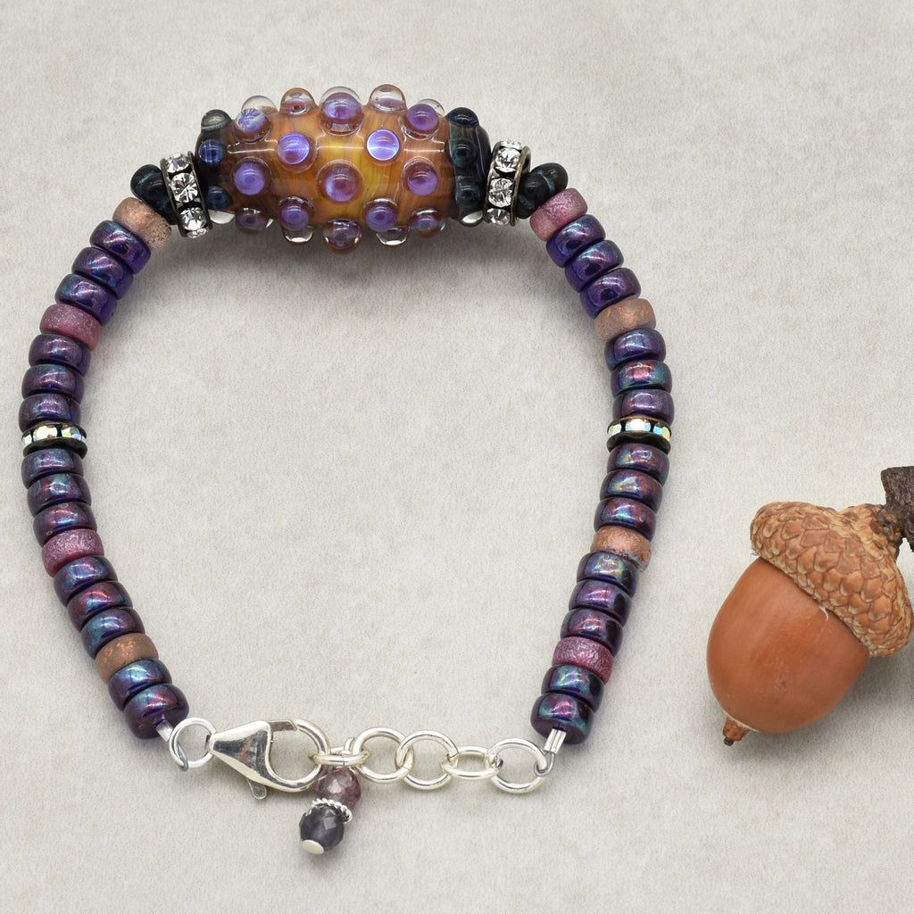 This image shows a bracelet. The jewelry piece is made of small round beads, with a larger central bead featuring purple and clear raised dots. There is also a clasp and extender chain visible, which is typical for a bracelet.