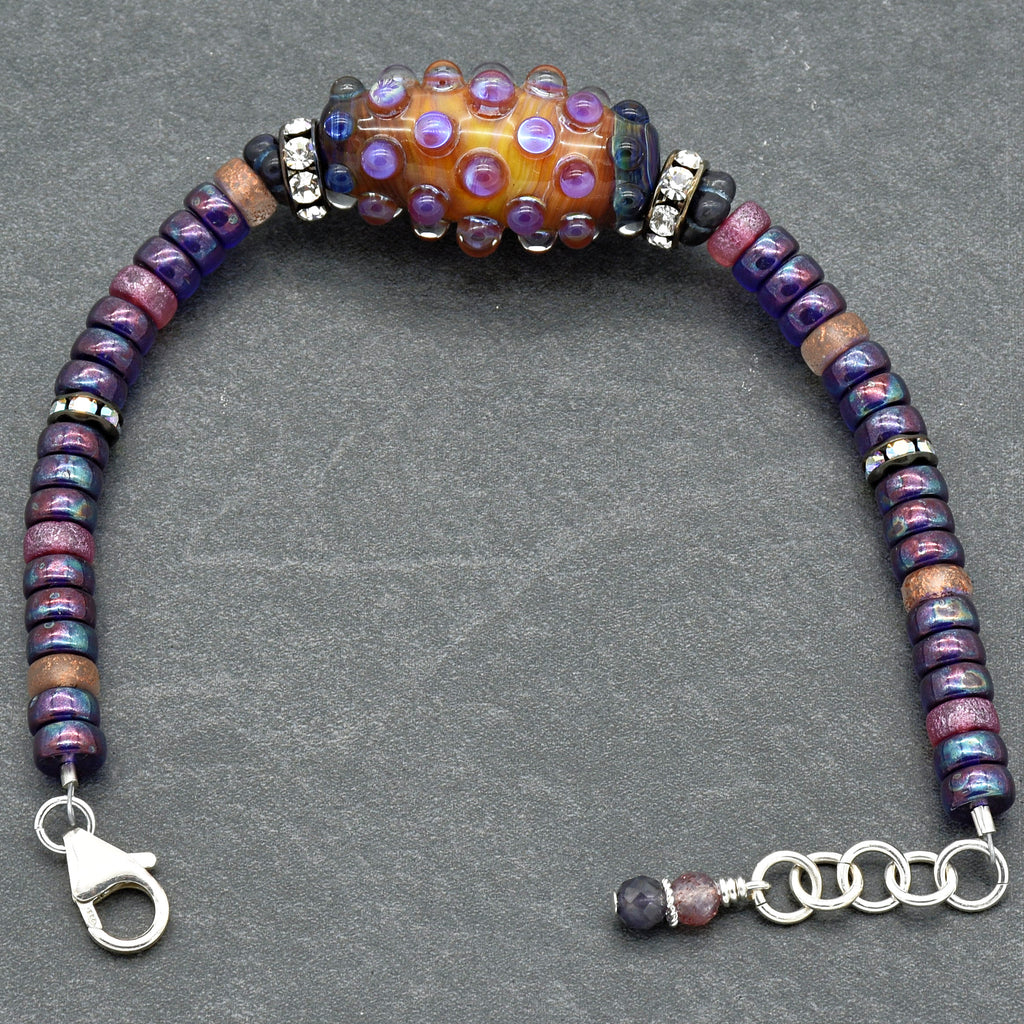 This image shows a bracelet. It features various beads in purple, blue, and pink tones along with a prominent central bead that has an orange and purple textured design. The bracelet also has a lobster claw clasp and an adjustable chain for sizing.