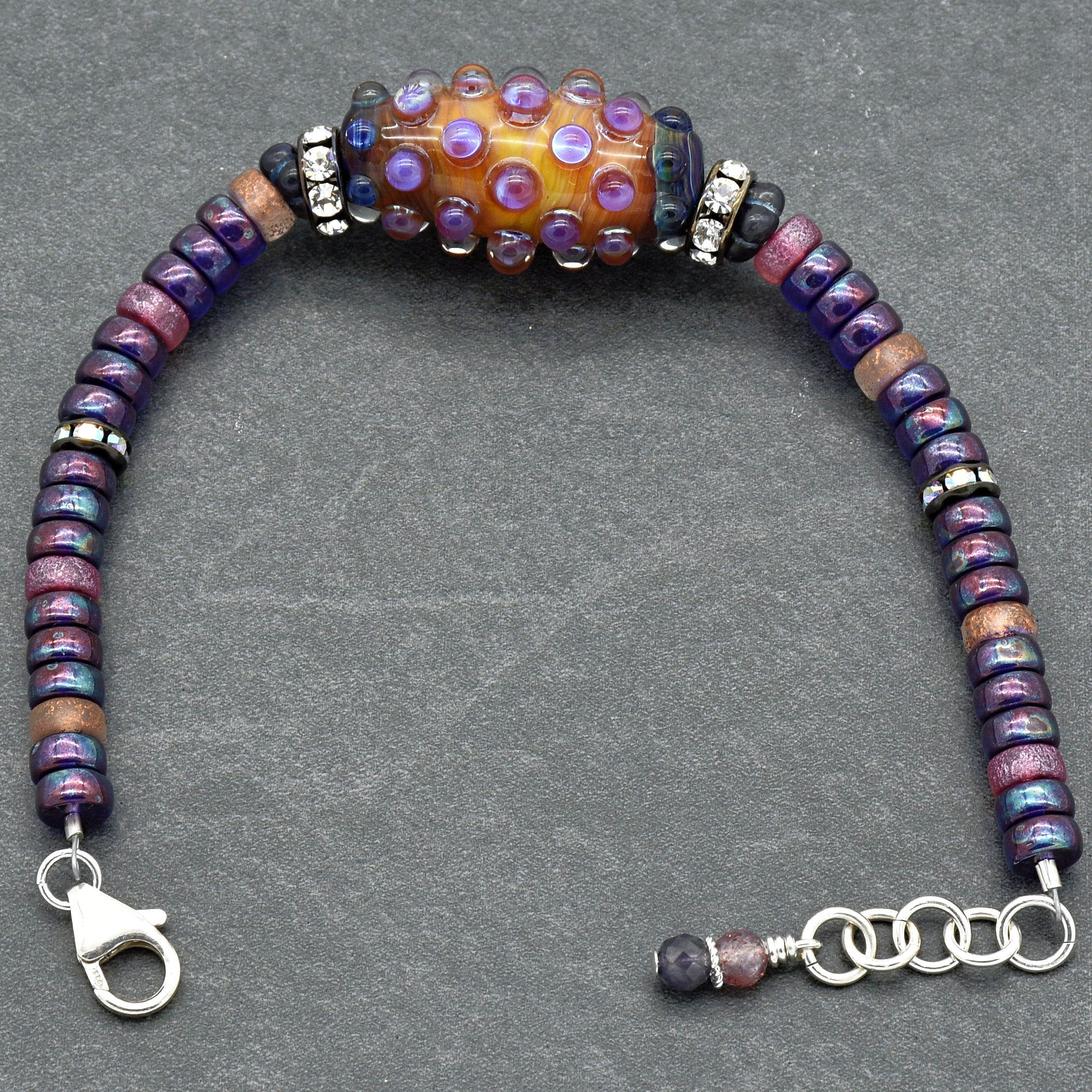 This image shows a bracelet. It features various beads in purple, blue, and pink tones along with a prominent central bead that has an orange and purple textured design. The bracelet also has a lobster claw clasp and an adjustable chain for sizing.