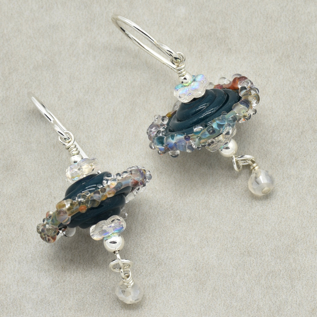 A pair of handmade silver wire-wrapped dangle earrings featuring dark blue glass beads, iridescent clear beads, and small white moonstone drops, displayed on a textured light gray background.