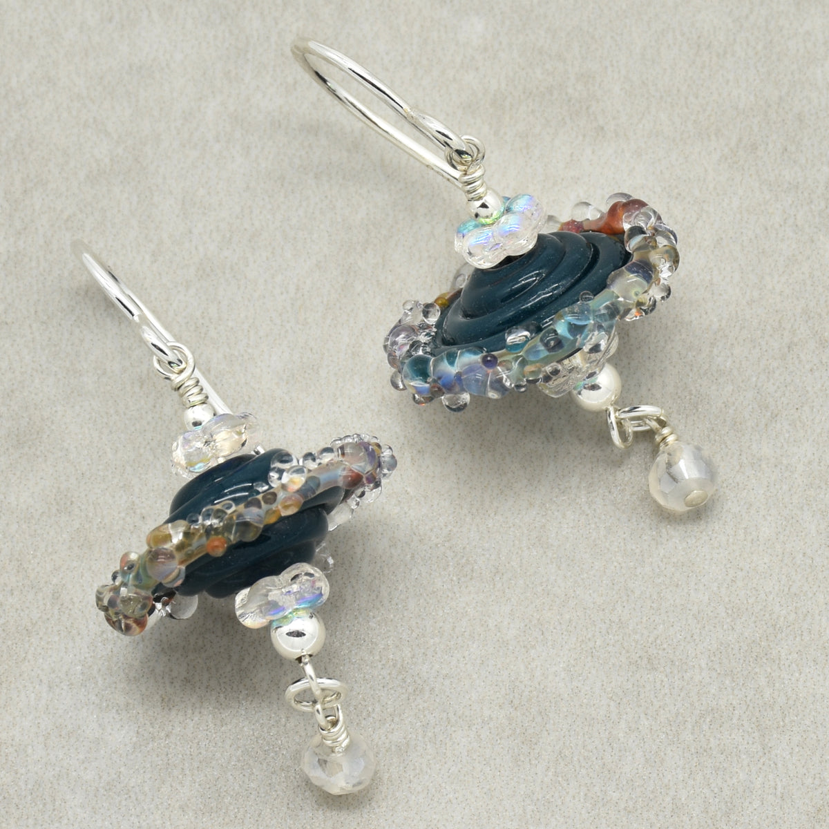 A pair of handmade silver wire-wrapped dangle earrings featuring dark blue glass beads, iridescent clear beads, and small white moonstone drops, displayed on a textured light gray background.