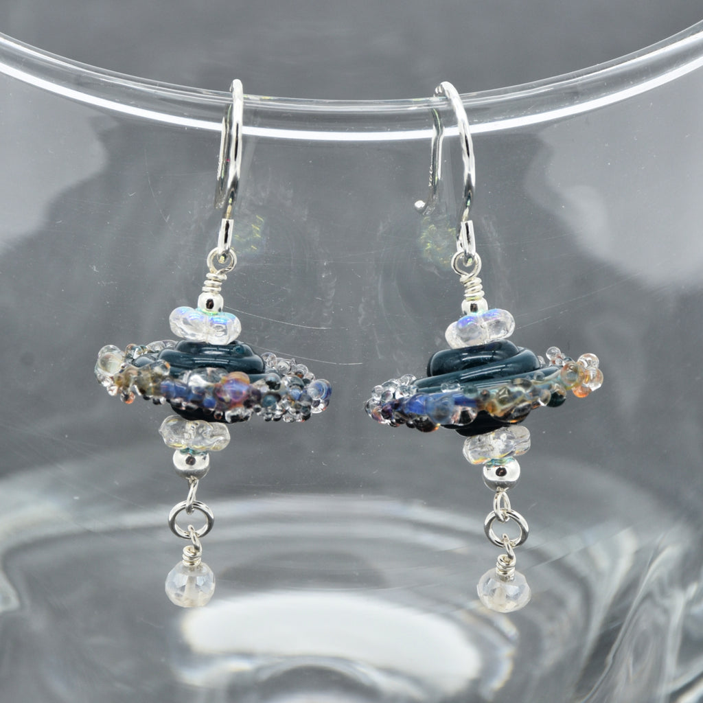 A pair of handmade dangle earrings with silver fishhooks, featuring dark blue lampwork glass beads, iridescent clear beads, and dangling clear faceted beads, displayed on a clear glass surface.