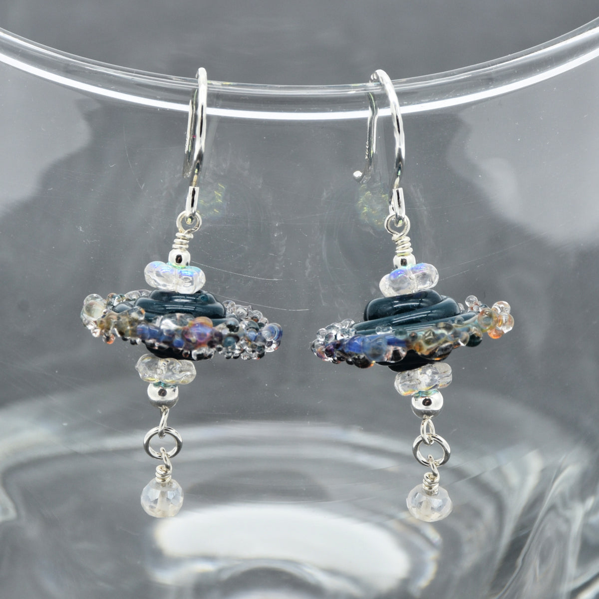 A pair of handmade dangle earrings with silver fishhooks, featuring dark blue lampwork glass beads, iridescent clear beads, and dangling clear faceted beads, displayed on a clear glass surface.
