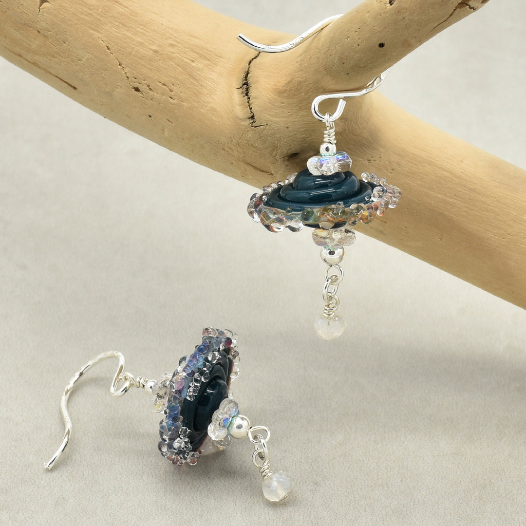 A pair of handmade dangle earrings with silver-tone French wire hooks, featuring dark blue glass beads, iridescent blue and purple beads, clear faceted beads, and rainbow moonstone beads on a branch.