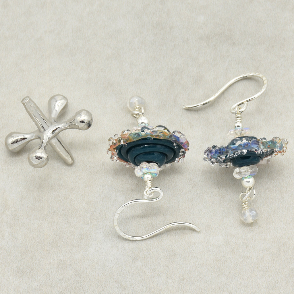 A pair of handmade earrings featuring a sterling silver ear wires, dark blue glass beads, iridescent accents, and small white beads, arranged on a textured light beige background.