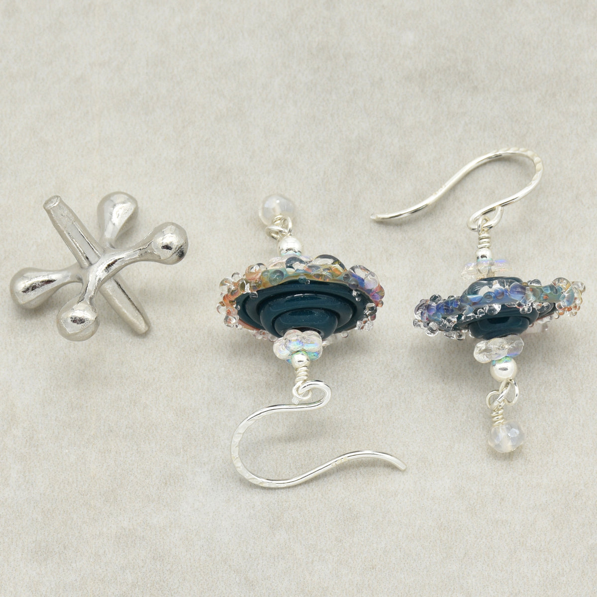 A pair of handmade earrings featuring a sterling silver ear wires, dark blue glass beads, iridescent accents, and small white beads, arranged on a textured light beige background.