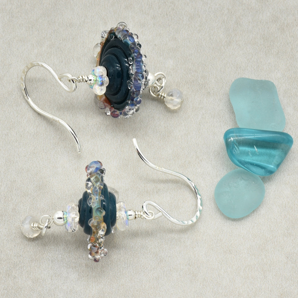 A pair of handmade silver dangle earrings with dark blue glass beads, clear faceted beads, and iridescent accents, displayed next to three light blue sea glass stones on a textured beige background.