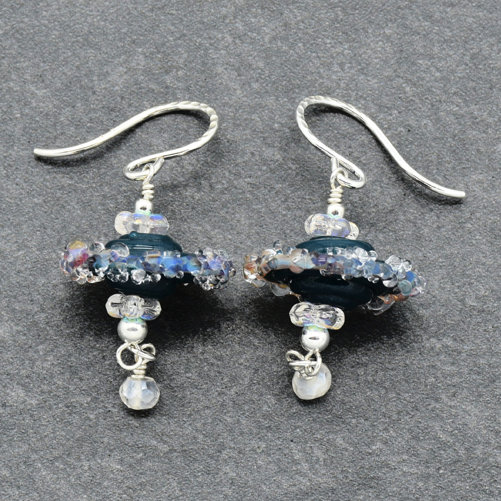 A pair of handmade dangle earrings with silver-tone French wire hooks, featuring dark blue glass beads, iridescent blue and purple beads, clear faceted beads, and rainbow moonstone beads on a dark gray textured background.