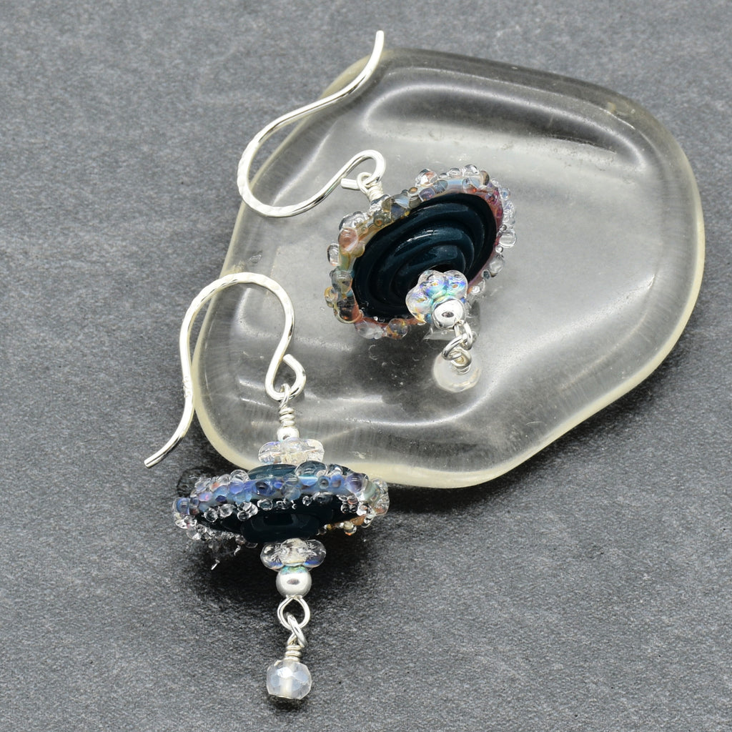 A pair of handmade dangle earrings with silver fishhook wires, featuring dark blue and black glass beads, clear faceted crystals, and small iridescent beads, displayed on a clear glass dish over a dark gray textured background.