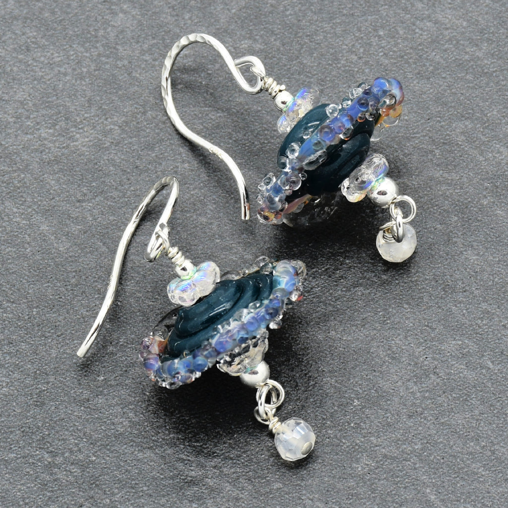 A pair of handmade dangle earrings featuring dark blue glass beads with iridescent blue and purple highlights, accented with clear faceted crystals and silver-toned metal hooks and beads, displayed on a dark gray textured background.
