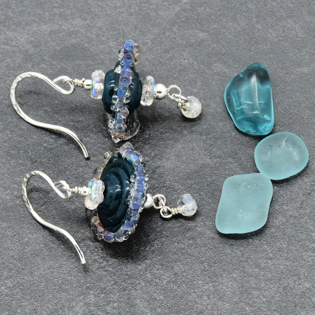 A pair of handmade dangle earrings with dark blue glass beads, iridescent rhinestones, and small clear beads, displayed next to three sea glass pieces on a dark gray textured background.