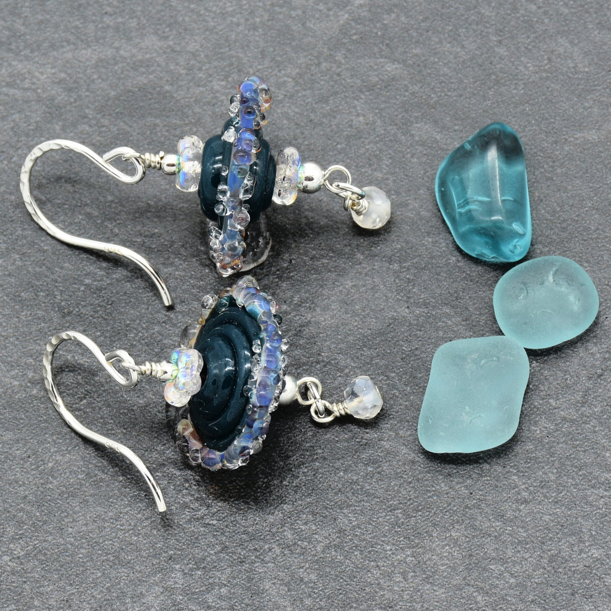 A pair of handmade dangle earrings with dark blue glass beads, iridescent rhinestones, and small clear beads, displayed next to three sea glass pieces on a dark gray textured background.