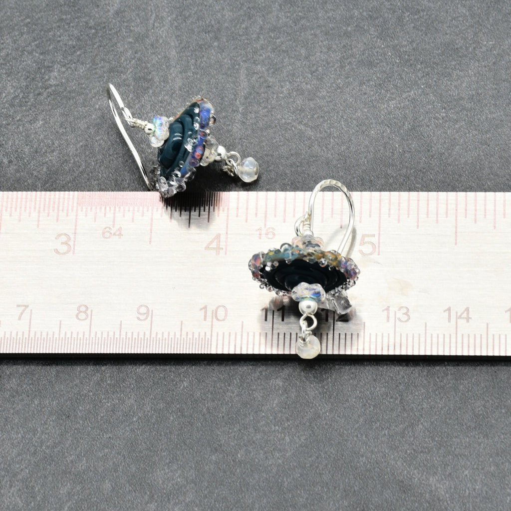 A pair of handmade silver earrings with dark blue lampwork glass beads and iridescent accents, shown next to a ruler for scale on a dark gray textured background.