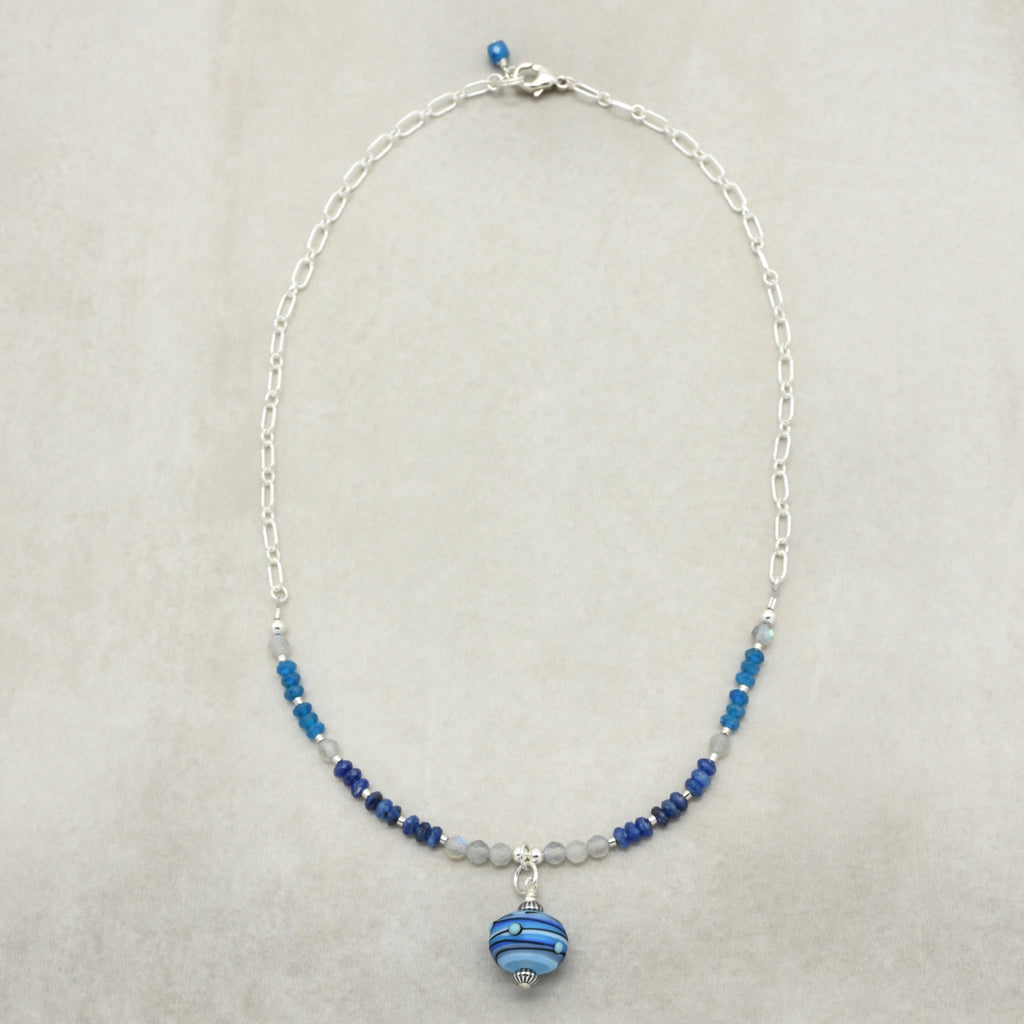 A delicate silver chain necklace featuring small blue and white beads, a silver-toned central charm, and a larger blue and white striped bead pendant, displayed on a neutral beige background.