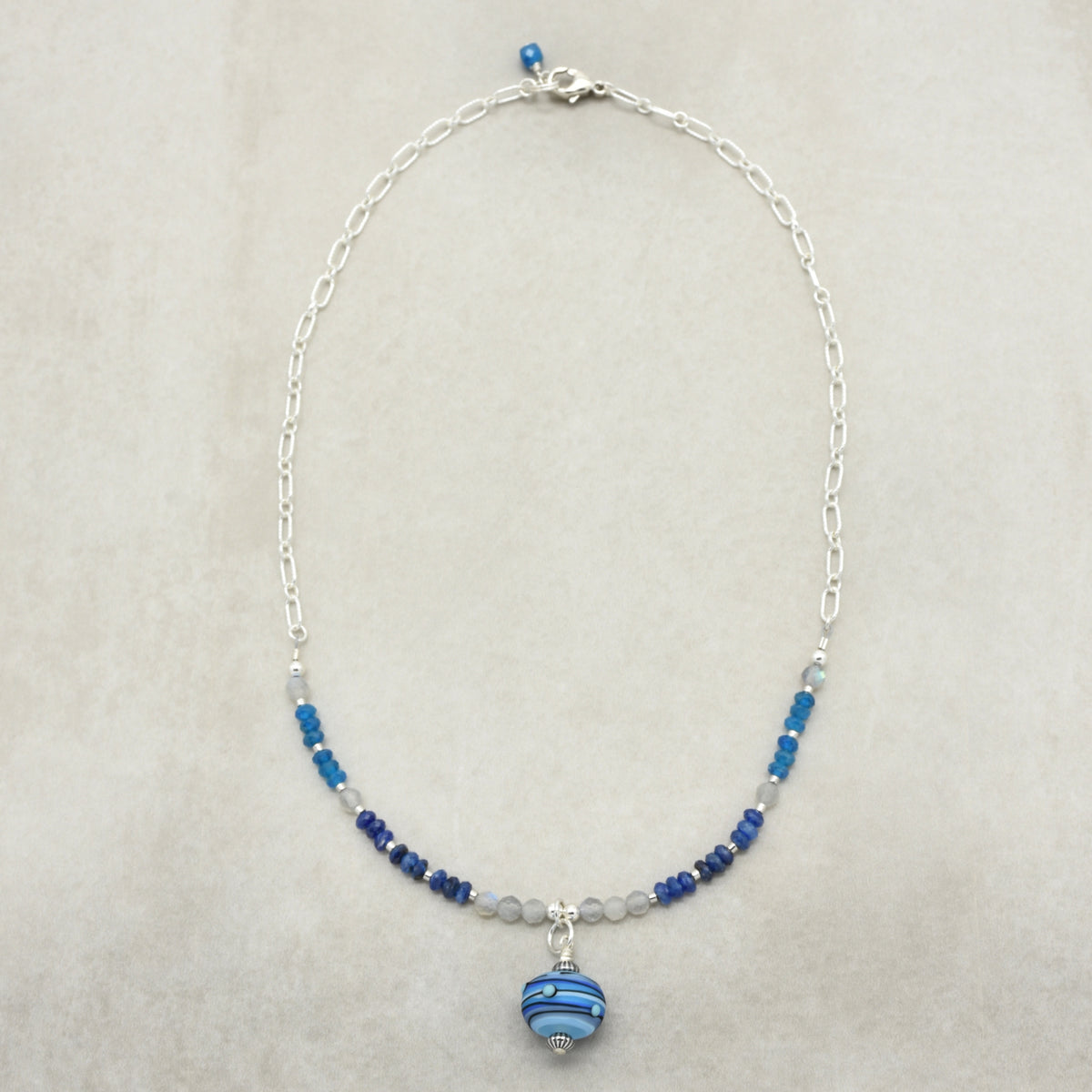 A delicate silver chain necklace featuring small blue and white beads, a silver-toned central charm, and a larger blue and white striped bead pendant, displayed on a neutral beige background.