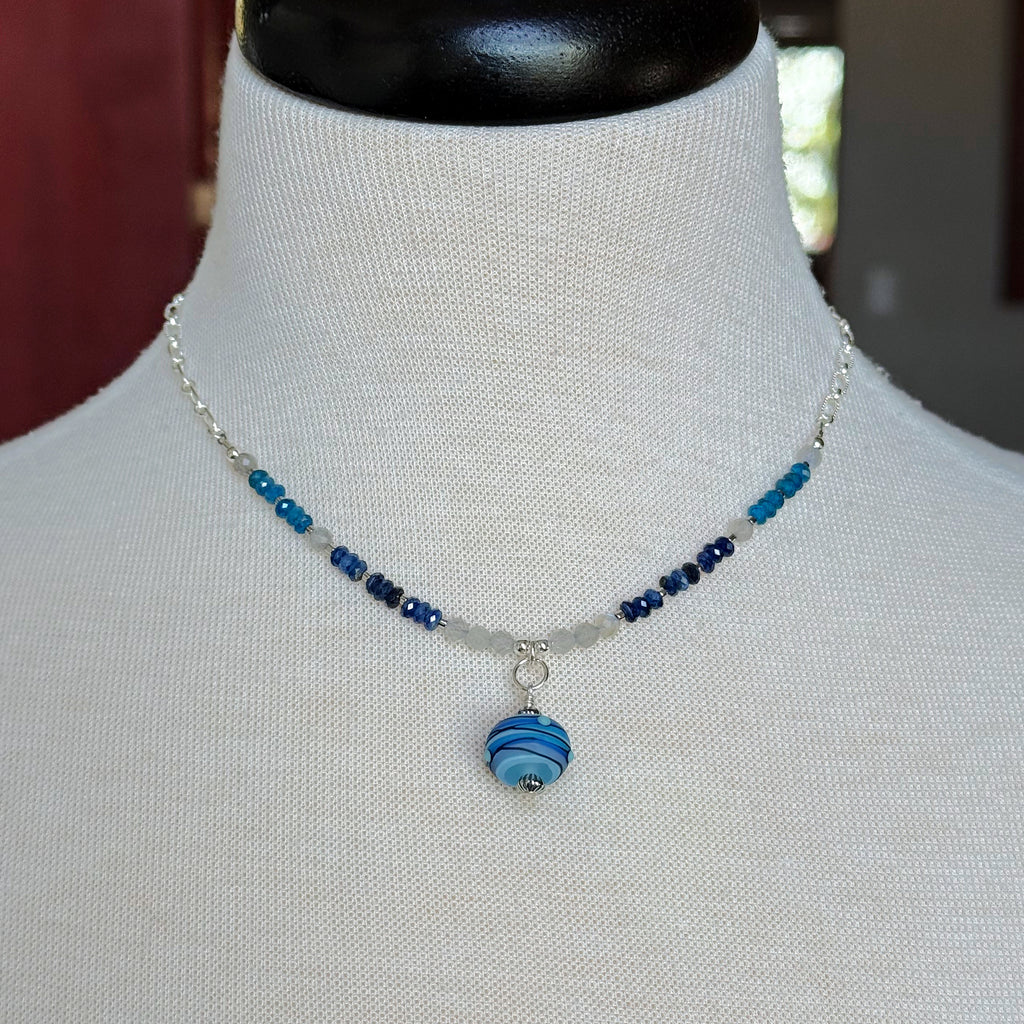 A beaded necklace with blue and navy beads and a striped blue glass pendant, displayed on a white fabric mannequin bust.
