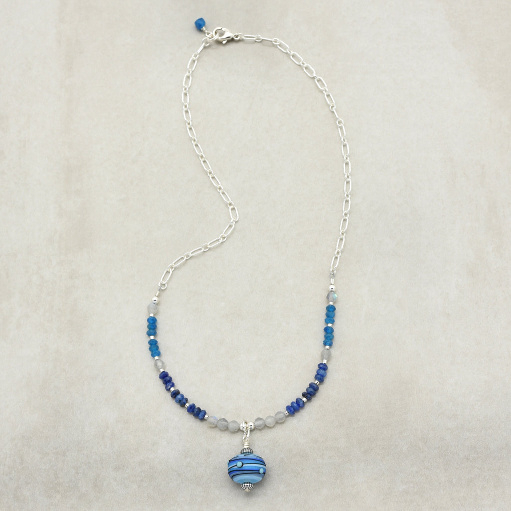 A silver chain necklace featuring blue and dark blue beads, clear faceted beads, and a striped blue glass pendant, displayed on a neutral beige background.