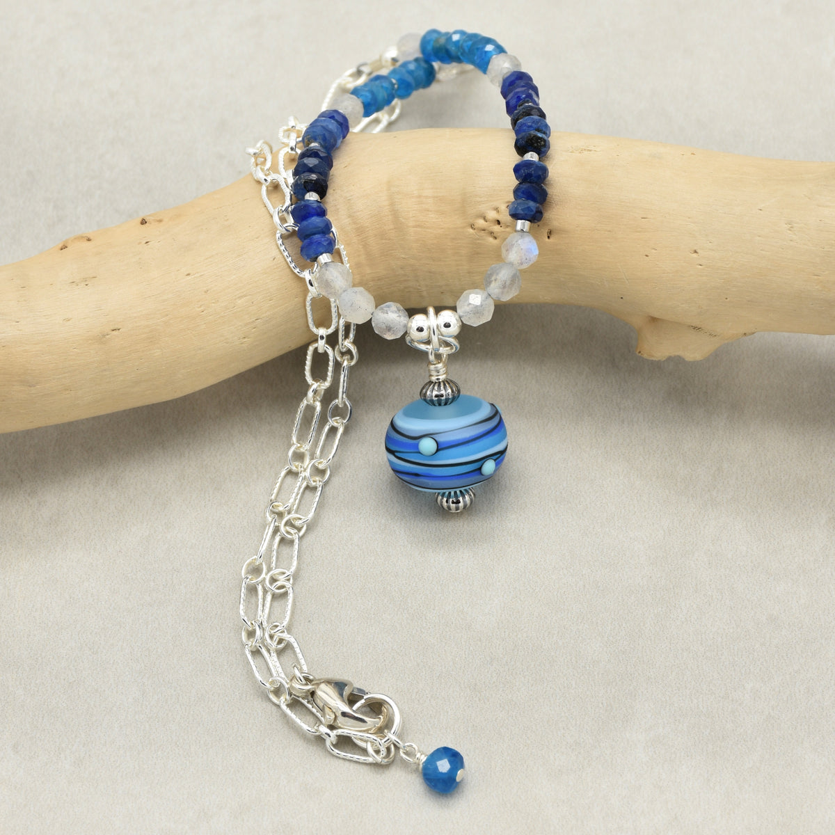 A beaded necklace with blue and white beads, a silver chain, and a round blue marbled glass charm, displayed on a light wood branch against a neutral background.