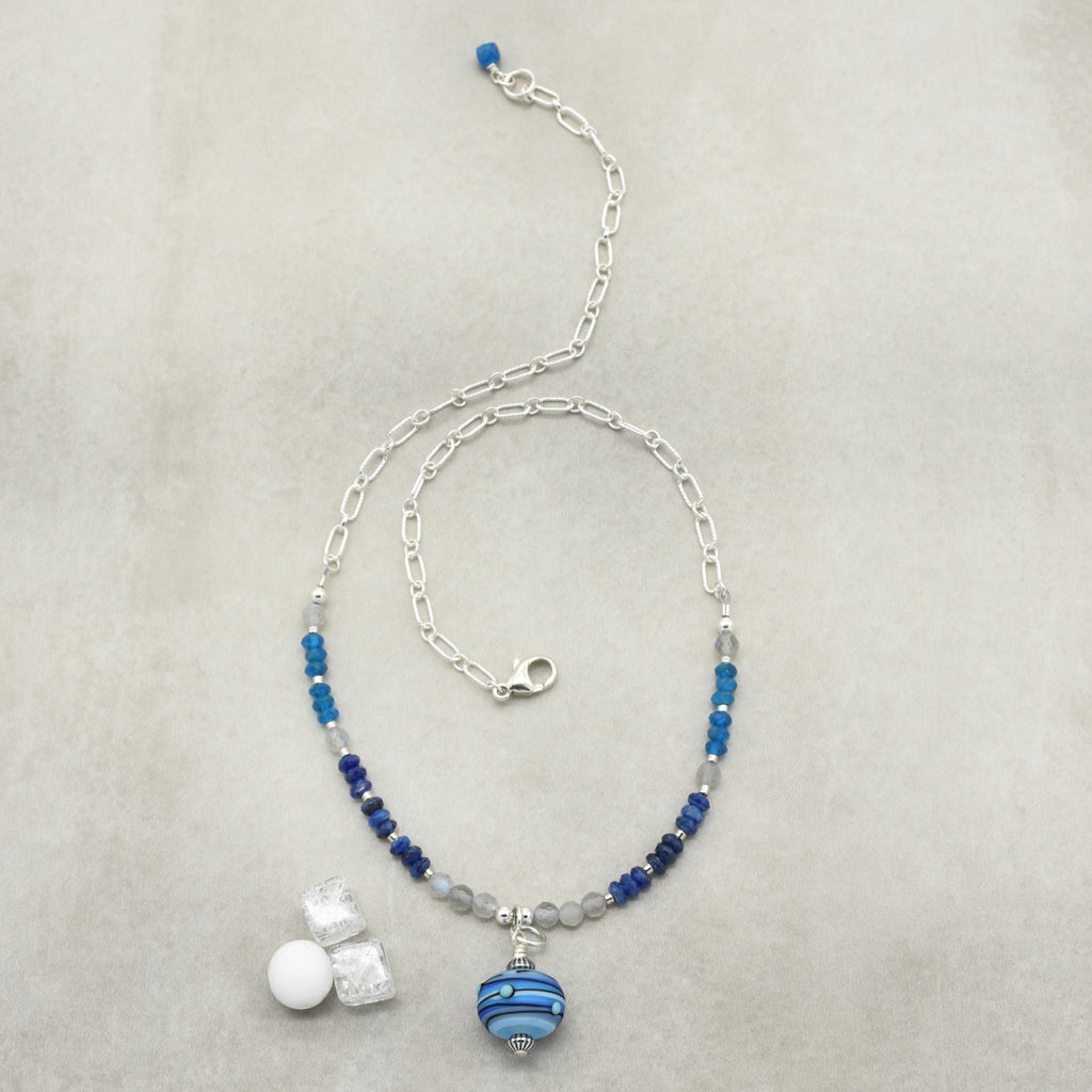 A beaded necklace with a silver paperclip chain, featuring blue and navy beads, gray translucent beads, and a blue and white striped glass bead pendant, on a textured light gray background.
