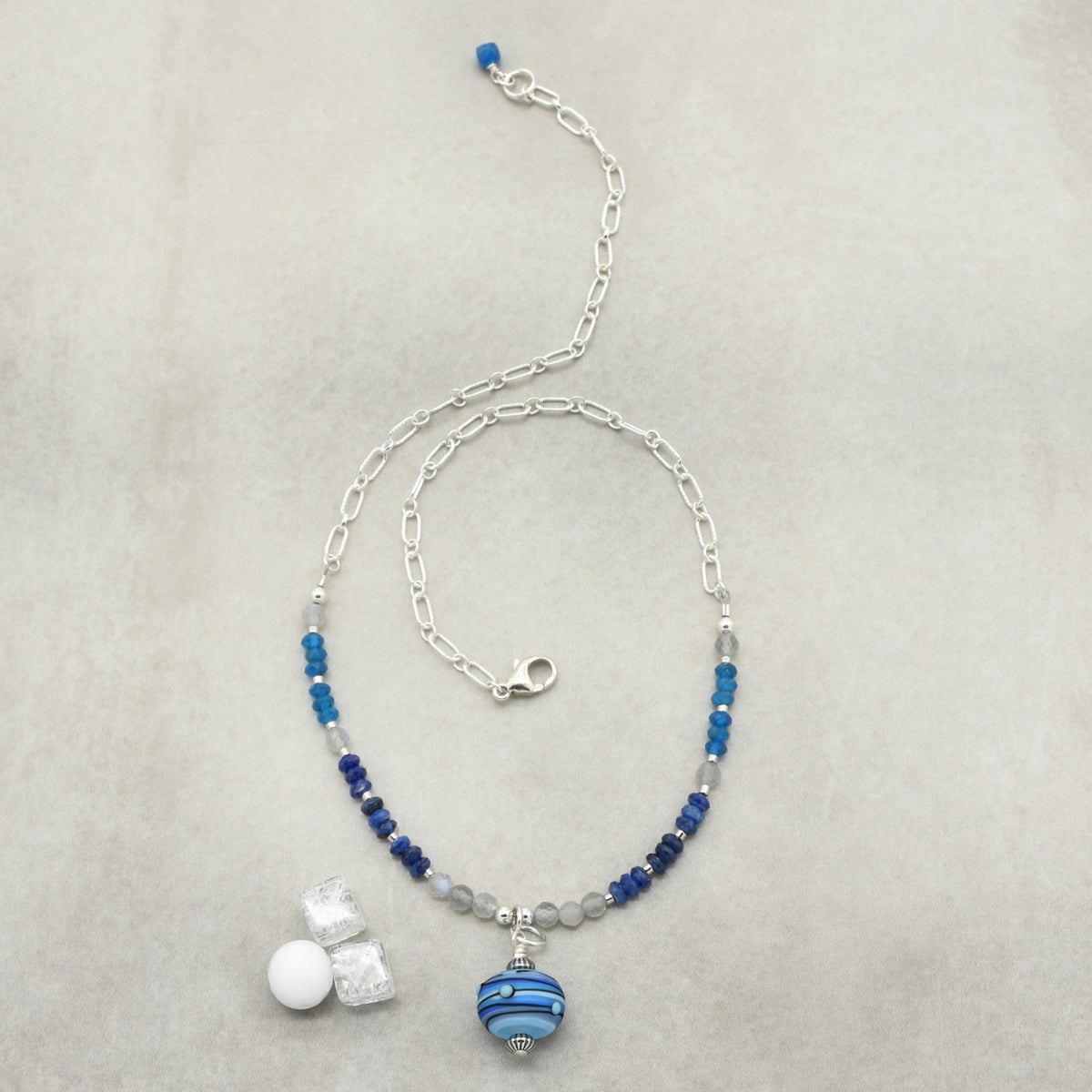 A beaded necklace with a silver paperclip chain, featuring blue and navy beads, gray translucent beads, and a blue and white striped glass bead pendant, on a textured light gray background.