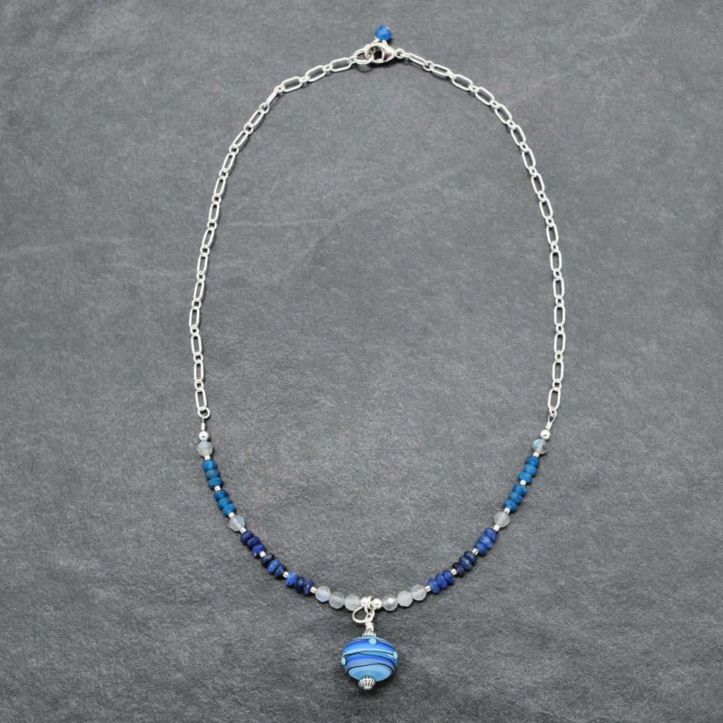 A silver chain necklace with a central cluster of blue and navy beads, a small silver bead, and a blue striped glass pendant, displayed on a textured gray background.