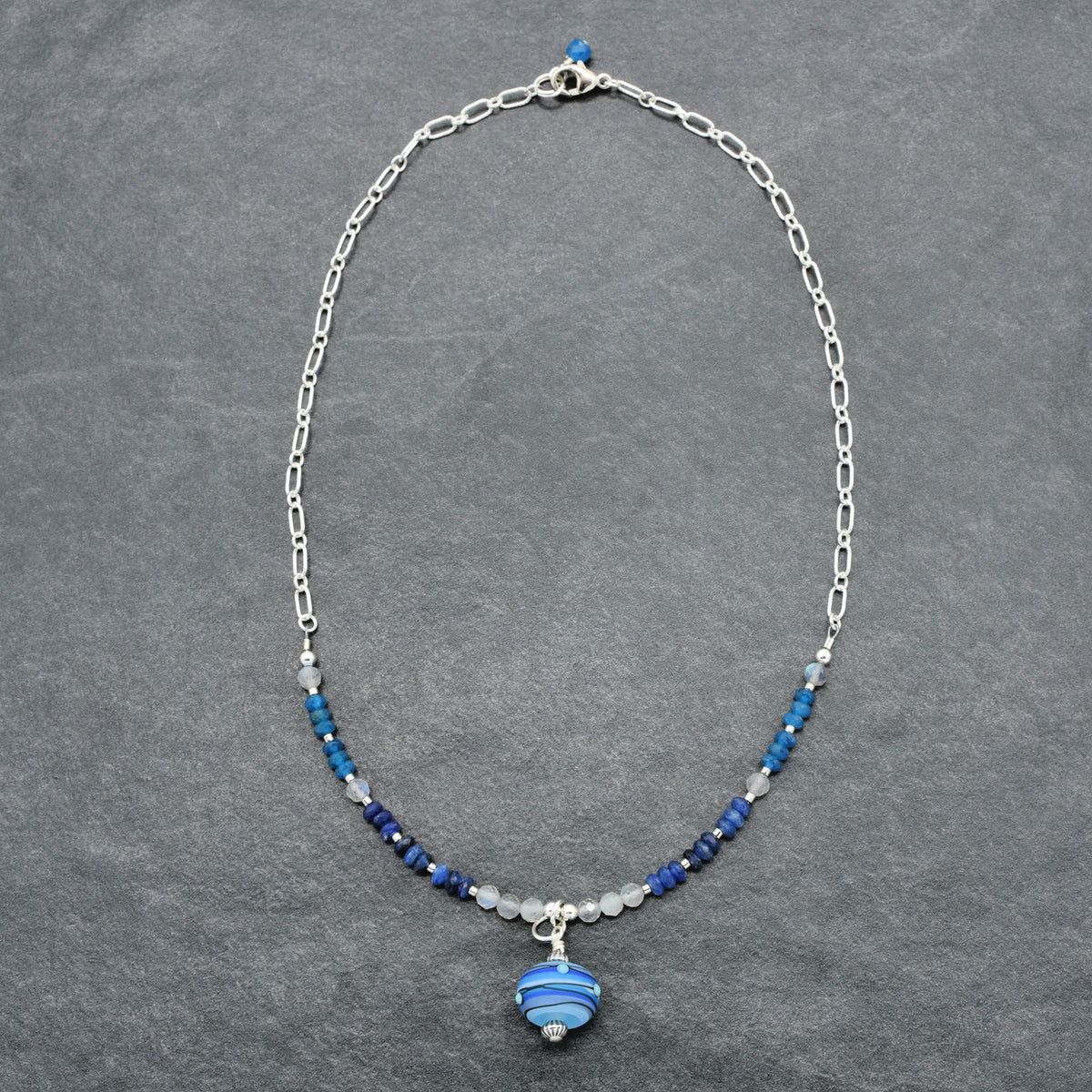 A silver chain necklace with a central cluster of blue and navy beads, a small silver bead, and a blue striped glass pendant, displayed on a textured gray background.