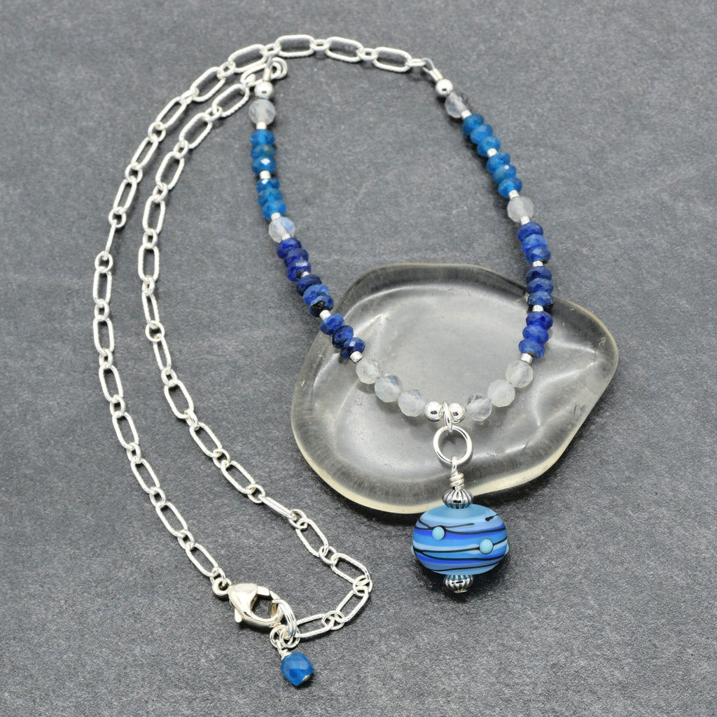 A silver chain necklace with blue and light gray beads and a blue striped glass pendant, displayed on a gray textured background with a clear stone prop.