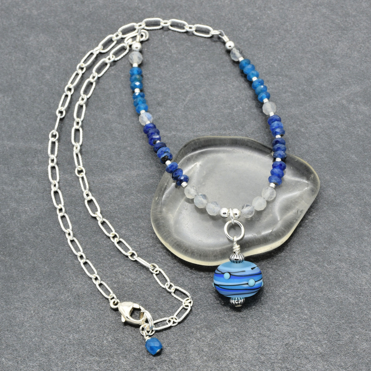 A silver chain necklace with blue and light gray beads and a blue striped glass pendant, displayed on a gray textured background with a clear stone prop.