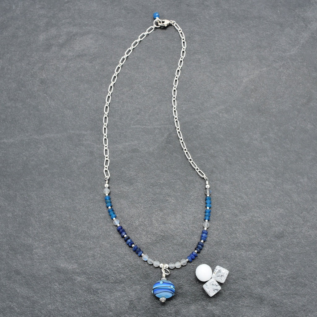 A silver chain necklace with blue and white beads, featuring a striped blue glass pendant displayed on a textured gray background.