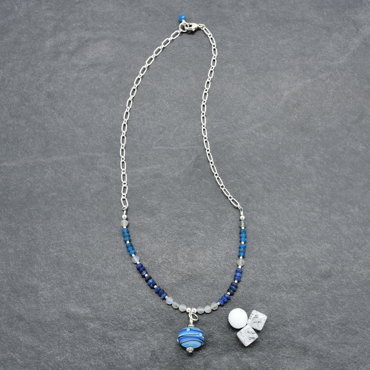 A silver chain necklace with blue and white beads, featuring a striped blue glass pendant displayed on a textured gray background.