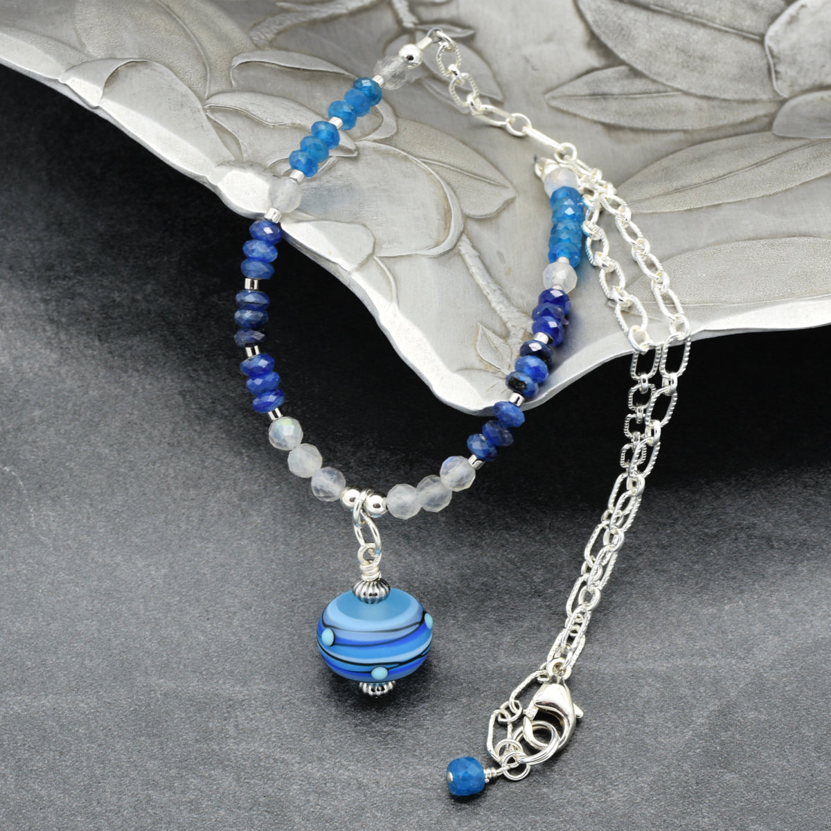 A beaded necklace with blue and white beads, a silver chain, and a round blue  bead pendant, displayed on a textured dark gray surface with a silver decorative plate in the background.