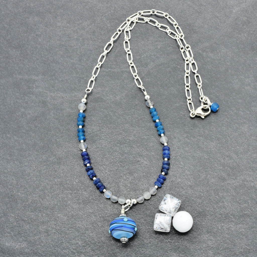 A silver chain necklace with blue beads, a blue and white striped glass bead pendant, displayed on a textured gray background.